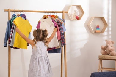 Photo of Little girl choosing clothes on rack in room