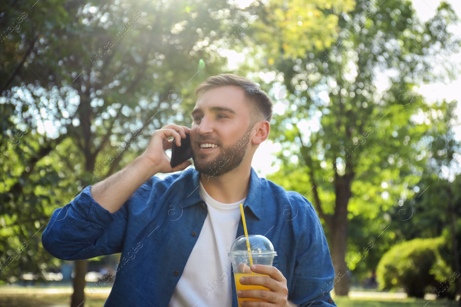 Young man with refreshing drink talking on smartphone in park Photo of Young man with refreshing drink talking on smartphone in park