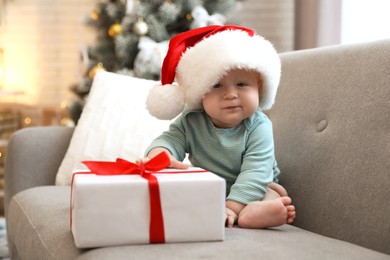 Cute baby in Santa hat with Christmas gift sitting on sofa at home Image of Cute baby in Santa hat with Christmas gift sitting on sofa at home