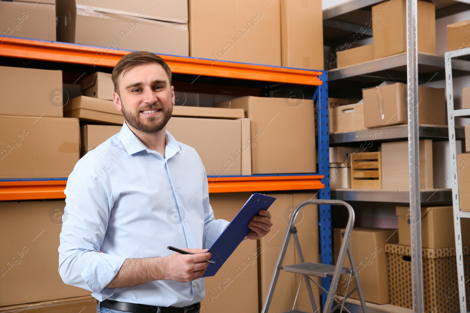 Young businessman with clipboard near rack of cardboard boxes at warehouse Photo of Young businessman with clipboard near rack of cardboard boxes at warehouse