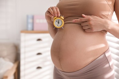Young pregnant woman pointing at alarm clock near her belly indoors, closeup. Time to give birth Photo of Young pregnant woman pointing at alarm clock near her belly indoors, closeup. Time to give birth
