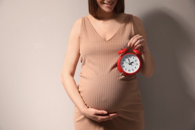 Young pregnant woman holding alarm clock near her belly on beige background, closeup. Time to give birth Photo of Young pregnant woman holding alarm clock near her belly on beige background, closeup. Time to give birth