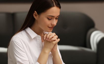 Religious young woman with clasped hands praying indoors Photo of Religious young woman with clasped hands praying indoors