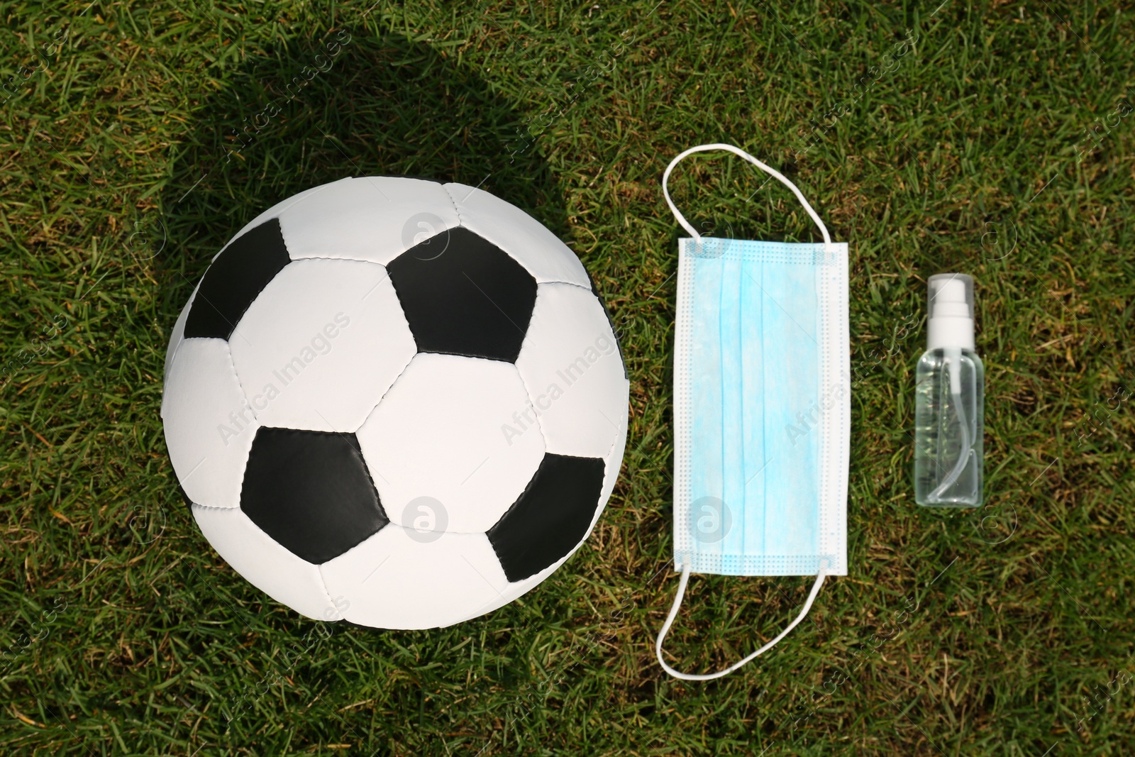 Photo of Football ball with hand sanitizer and protective mask on green grass, flat lay