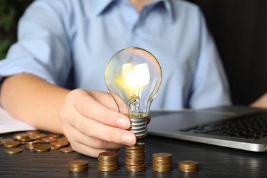 Woman with light bulb and coins at black wooden table, closeup. Power saving Photo of Woman with light bulb and coins at black wooden table, closeup. Power saving