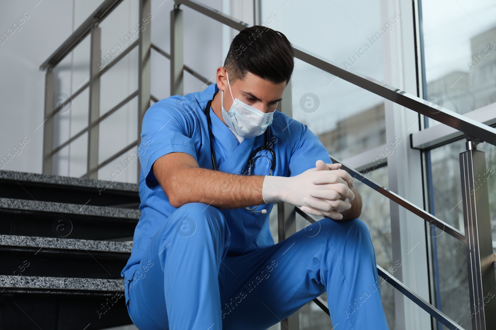 Sad doctor in facial mask on stairs indoors. Stress of health care workers during coronavirus pandemic Photo of Sad doctor in facial mask on stairs indoors. Stress of health care workers during coronavirus pandemic
