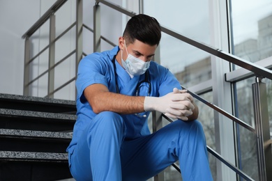 Sad doctor in facial mask on stairs indoors. Stress of health care workers during coronavirus pandemic Photo of Sad doctor in facial mask on stairs indoors. Stress of health care workers during coronavirus pandemic