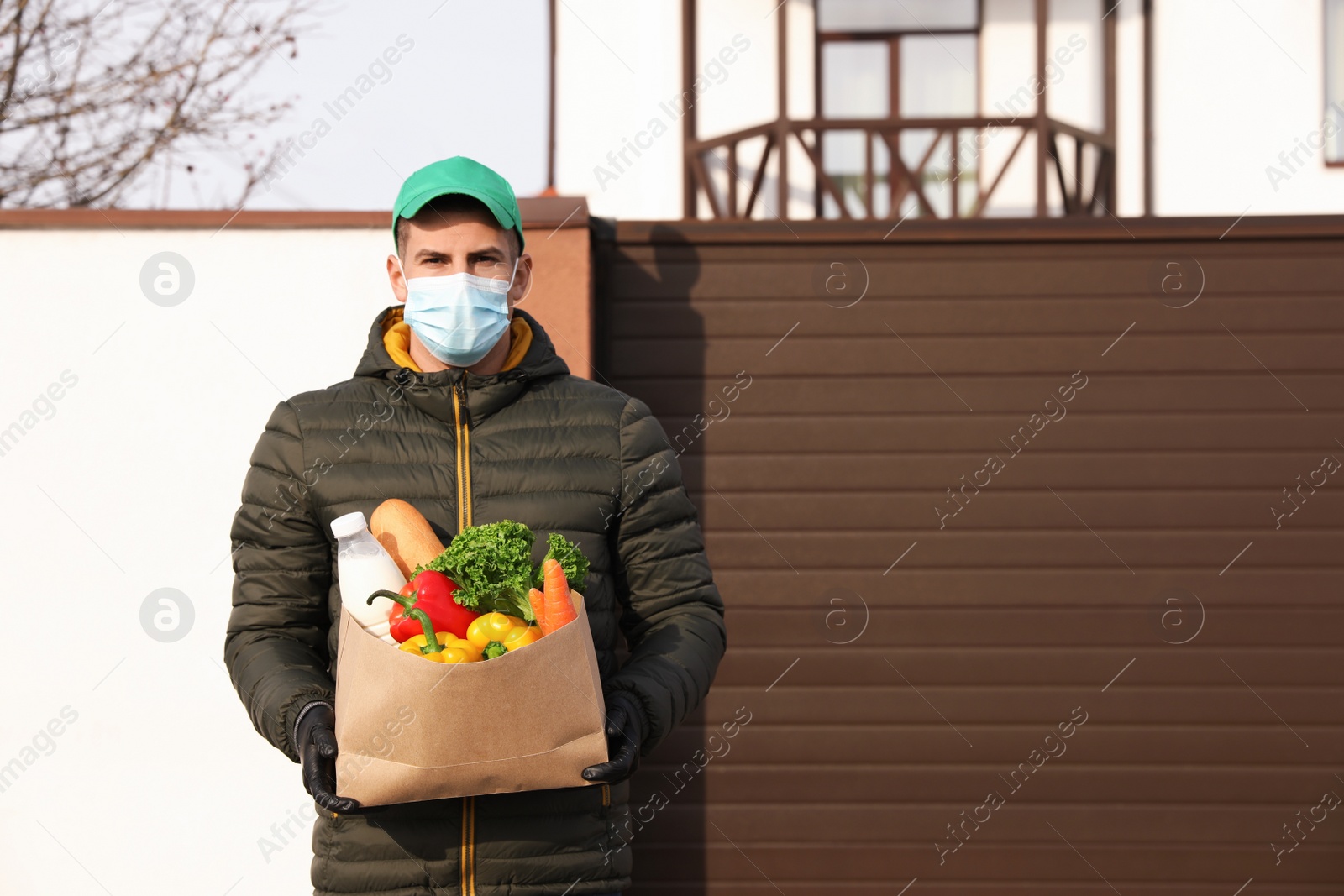 Photo of Courier in medical mask holding paper bag with groceries near house outdoors. Delivery service during quarantine due to Covid-19 outbreak