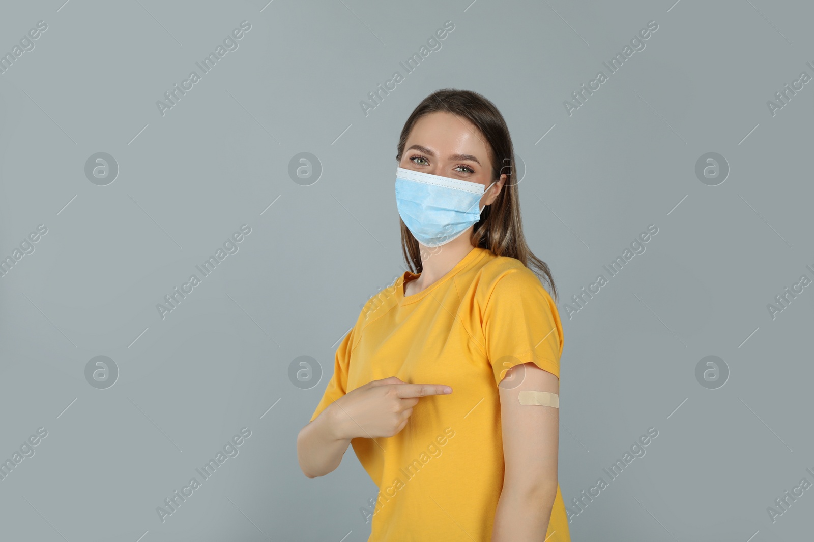 Vaccinated woman with protective mask showing medical plaster on her arm against grey background Photo of Vaccinated woman with protective mask showing medical plaster on her arm against grey background