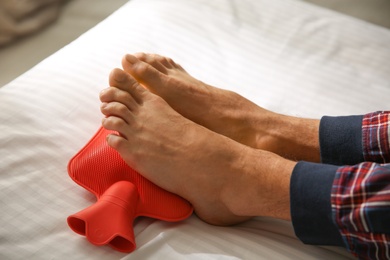 Man warming feet with hot water bottle on bed, closeup Photo of Man warming feet with hot water bottle on bed, closeup