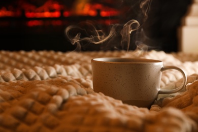 Cup of hot drink on knitted plaid near fireplace indoors. Space for text Photo of Cup of hot drink on knitted plaid near fireplace indoors. Space for text