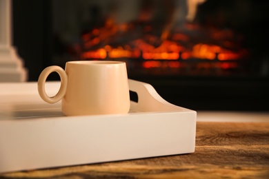 Cup with hot drink in tray on wooden table against fireplace, space for text Photo of Cup with hot drink in tray on wooden table against fireplace, space for text