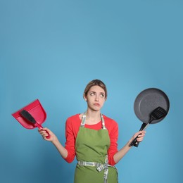 Young housewife with dustpan, brush, frying pan and spatula on light blue background Photo of Young housewife with dustpan, brush, frying pan and spatula on light blue background