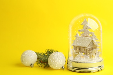 Beautiful snow globe, Christmas balls and fir branch on yellow background, space for text Photo of Beautiful snow globe, Christmas balls and fir branch on yellow background, space for text
