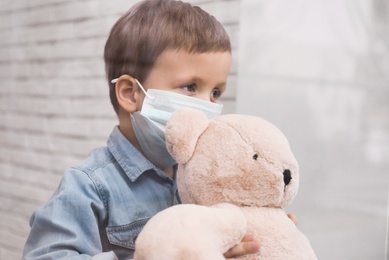 Sad little boy in protective mask looking out of window indoors, view from outside. Staying at home during coronavirus pandemic Photo of Sad little boy in protective mask looking out of window indoors, view from outside. Staying at home during coronavirus pandemic