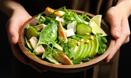 Woman holding bowl of delicious salad with chicken, arugula and avocado on dark background, closeup Photo of Woman holding bowl of delicious salad with chicken, arugula and avocado on dark background, closeup