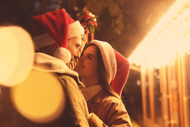 Happy couple in Santa hats standing under mistletoe bunch outdoors, bokeh effect Photo of Happy couple in Santa hats standing under mistletoe bunch outdoors, bokeh effect