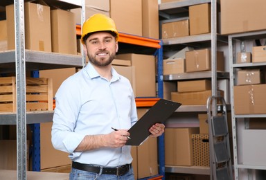 Young man with clipboard near rack of cardboard boxes at warehouse Photo of Young man with clipboard near rack of cardboard boxes at warehouse