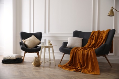 Comfortable sofa with knitted blanket, armchair and coffee table in stylish room interior Photo of Comfortable sofa with knitted blanket, armchair and coffee table in stylish room interior