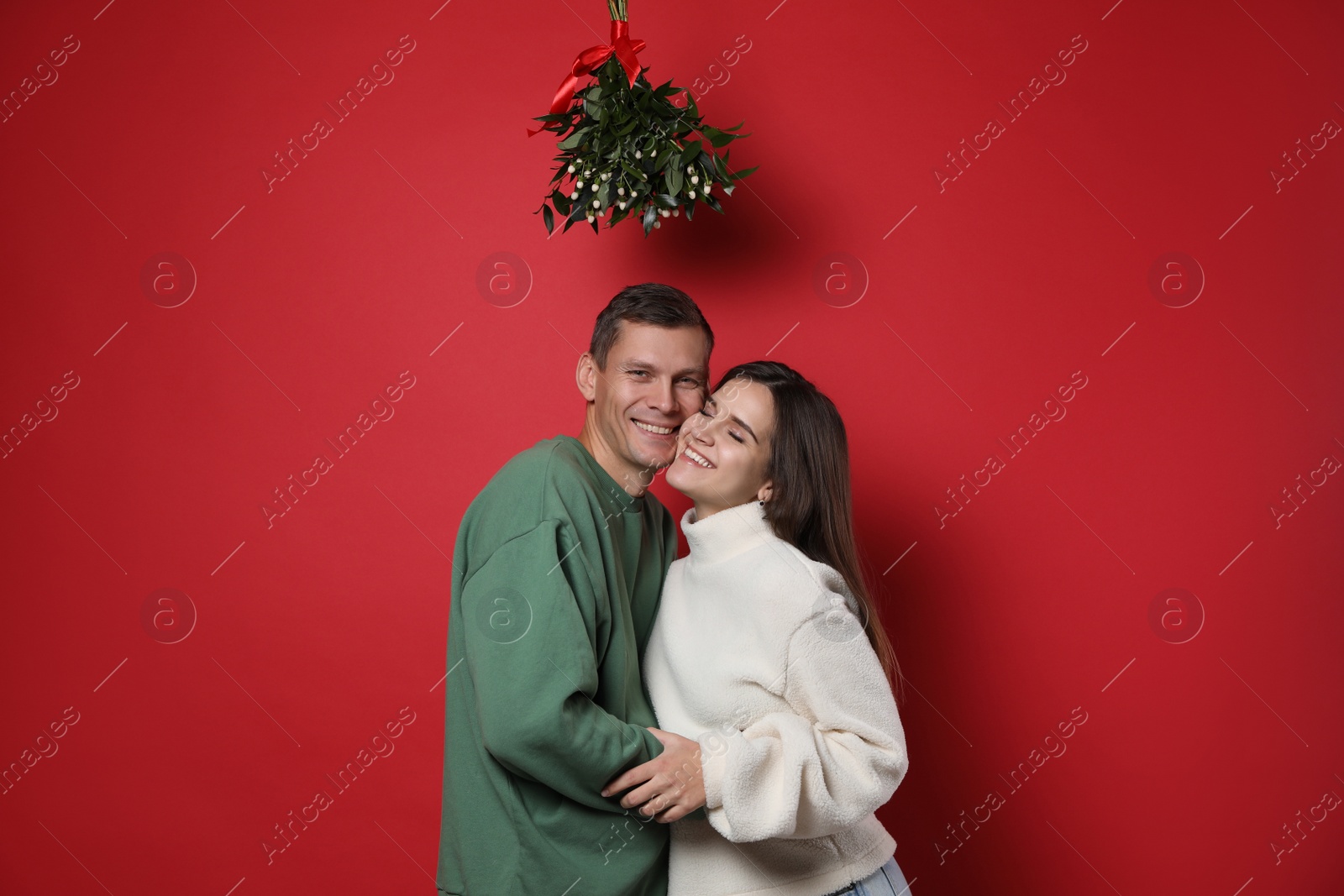 Happy couple standing under mistletoe bunch on red background Photo of Happy couple standing under mistletoe bunch on red background