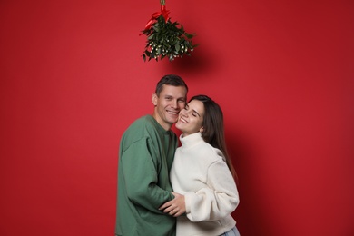 Happy couple standing under mistletoe bunch on red background Photo of Happy couple standing under mistletoe bunch on red background