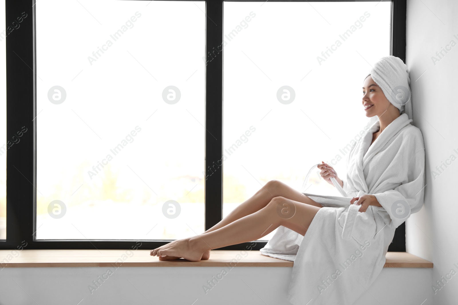 Young woman with towel wearing bathrobe near window Photo of Young woman with towel wearing bathrobe near window
