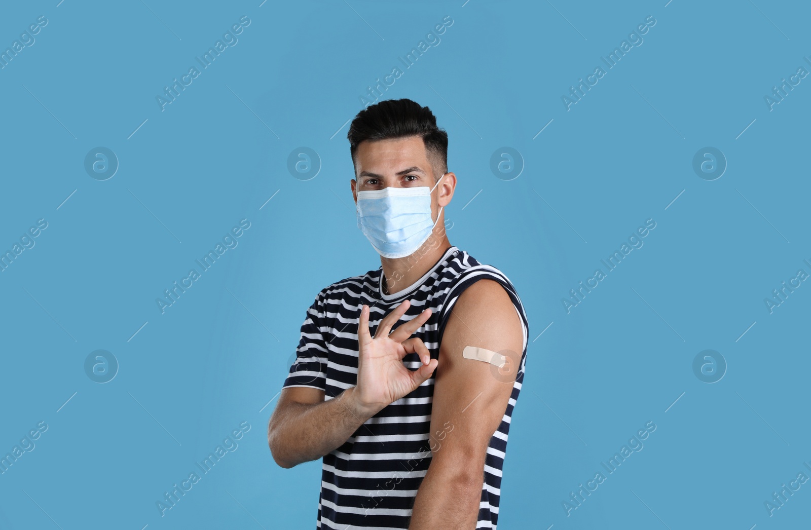 Vaccinated man with protective mask and medical plaster on his arm showing okay gesture against light blue background Photo of Vaccinated man with protective mask and medical plaster on his arm showing okay gesture against light blue background