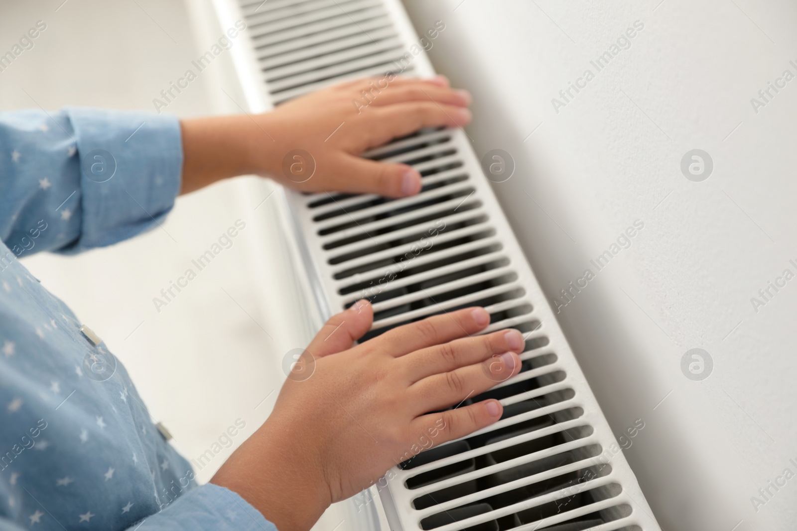 Child warming hands on heating radiator near white wall, closeup Photo of Child warming hands on heating radiator near white wall, closeup