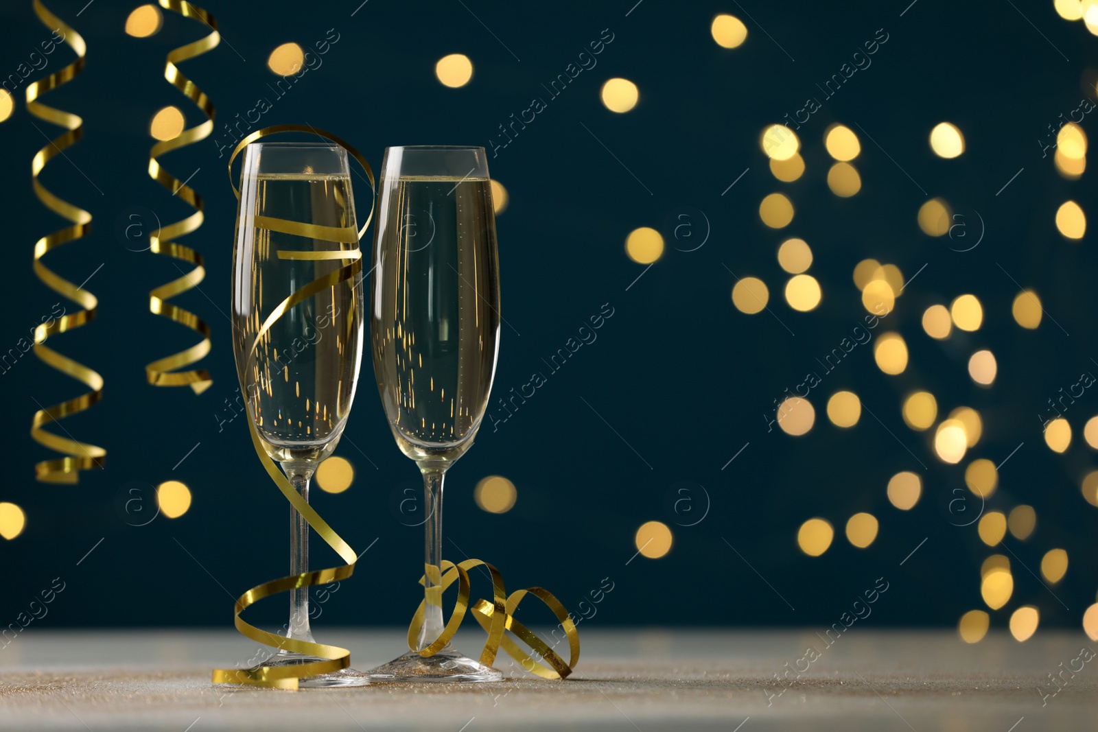 Photo of Glasses of champagne and serpentine streamers on table against dark blue background with blurred lights. Space for text