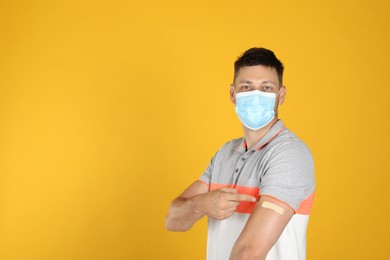 Vaccinated man with protective mask showing medical plaster on his arm against yellow background. Space for text Photo of Vaccinated man with protective mask showing medical plaster on his arm against yellow background. Space for text
