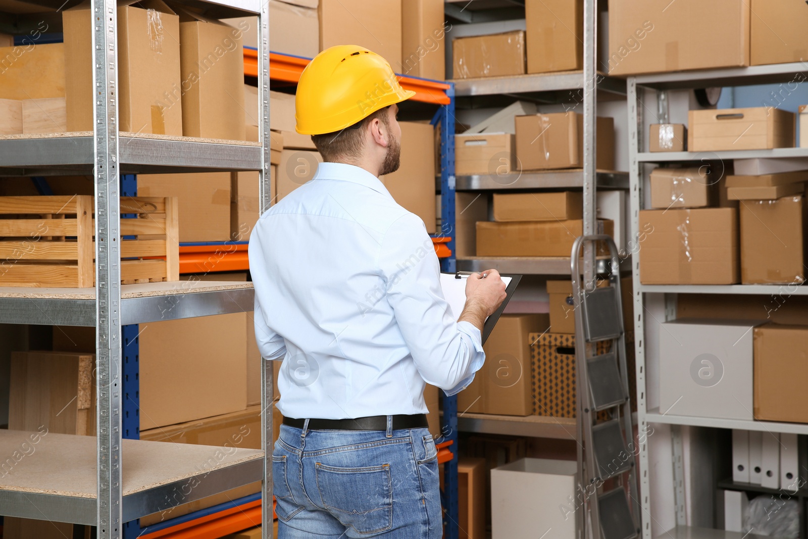Young man with clipboard near rack of cardboard boxes at warehouse, back view Photo of Young man with clipboard near rack of cardboard boxes at warehouse, back view