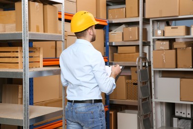 Young man with clipboard near rack of cardboard boxes at warehouse, back view Photo of Young man with clipboard near rack of cardboard boxes at warehouse, back view