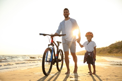 Photo of Happy father and son with bicycle on sandy beach near sea at sunset