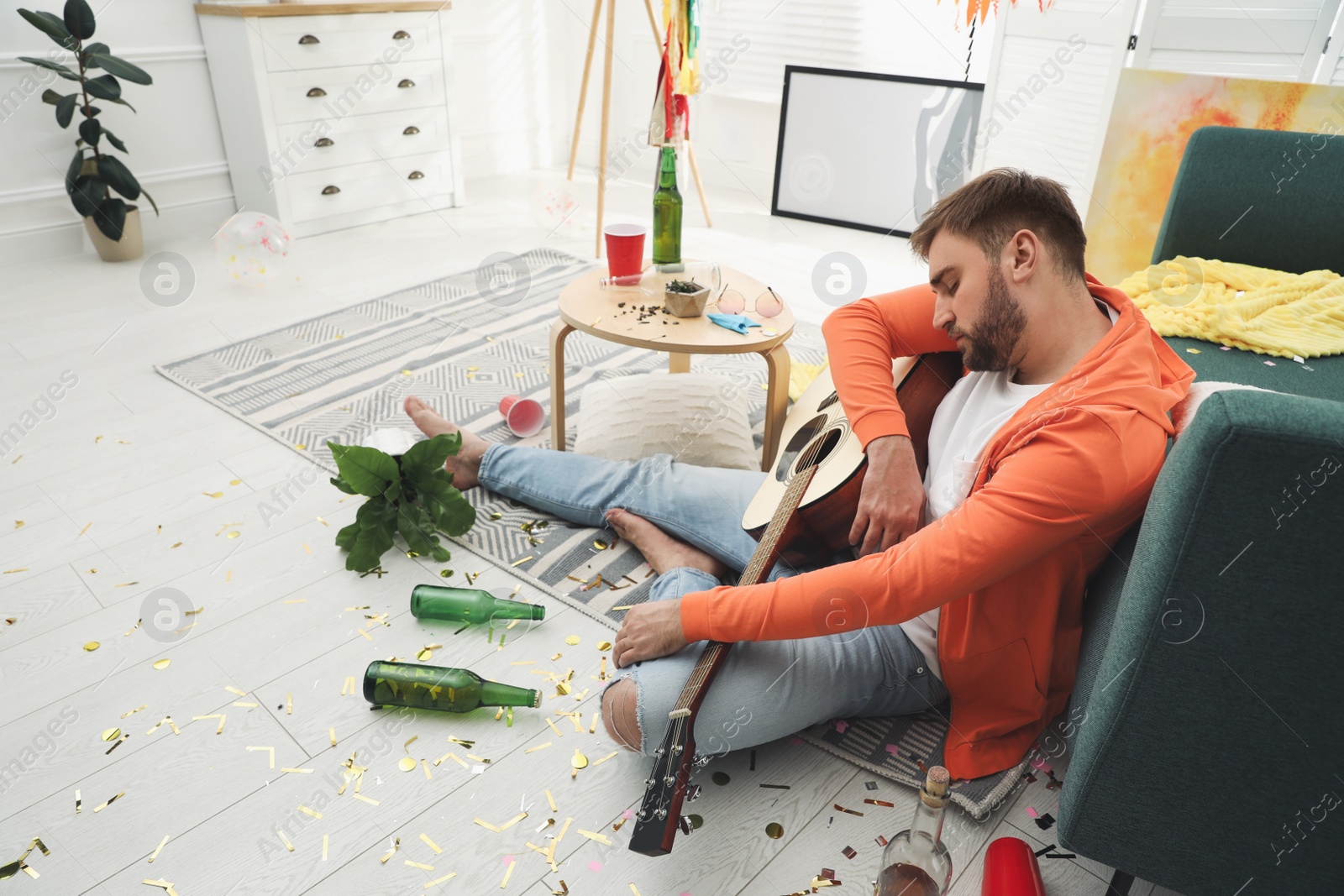 Young man with guitar sleeping near sofa in messy room after party Photo of Young man with guitar sleeping near sofa in messy room after party