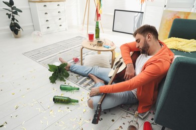 Young man with guitar sleeping near sofa in messy room after party Photo of Young man with guitar sleeping near sofa in messy room after party