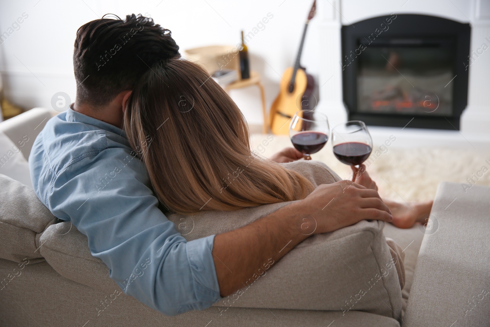 Couple with glasses of wine resting on sofa near fireplace at home, back view Photo of Couple with glasses of wine resting on sofa near fireplace at home, back view