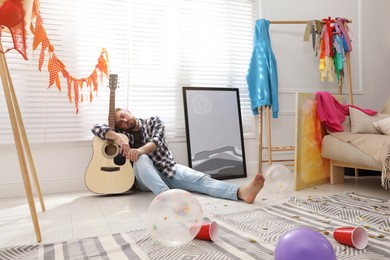 Young man with guitar sleeping near window in messy room after party Photo of Young man with guitar sleeping near window in messy room after party