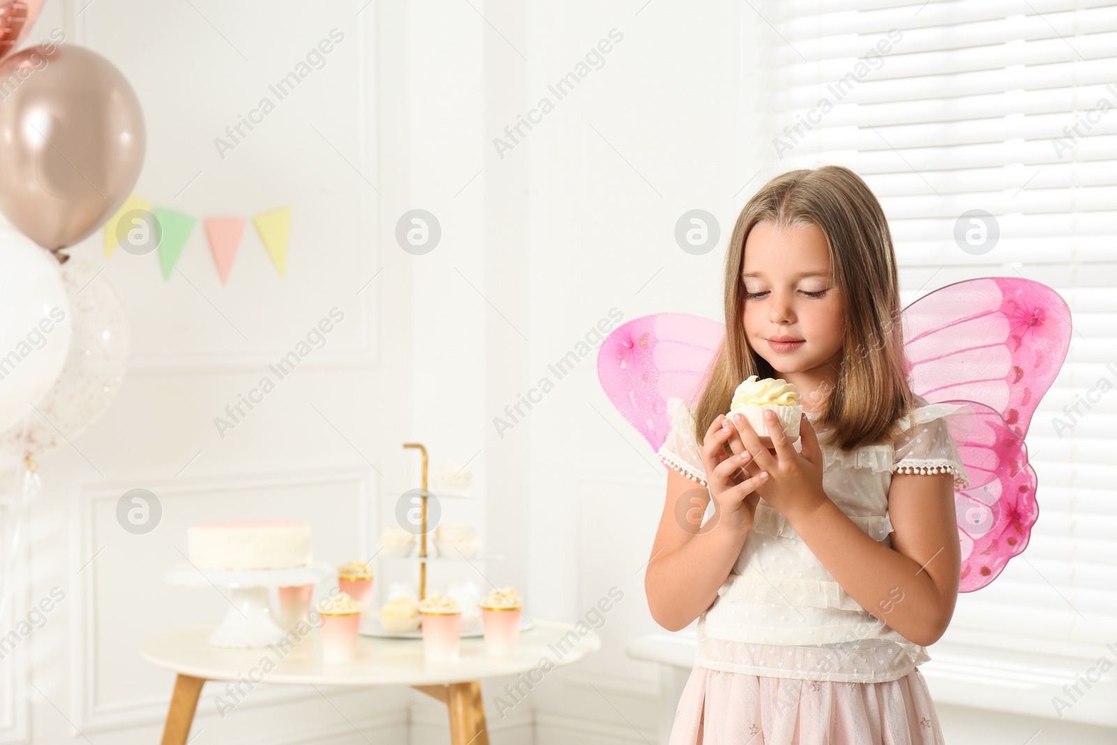 Cute little girl wearing fairy costume with delicious cupcake in decorated room. Space for text Photo of Cute little girl wearing fairy costume with delicious cupcake in decorated room. Space for text