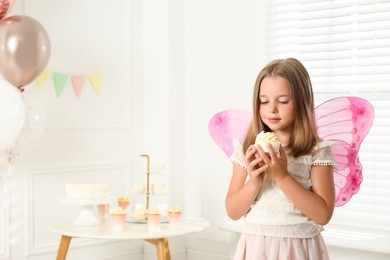 Cute little girl wearing fairy costume with delicious cupcake in decorated room. Space for text Photo of Cute little girl wearing fairy costume with delicious cupcake in decorated room. Space for text