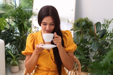 Beautiful woman with cup sitting in wicker armchair near houseplants indoors. Interior design Photo of Beautiful woman with cup sitting in wicker armchair near houseplants indoors. Interior design