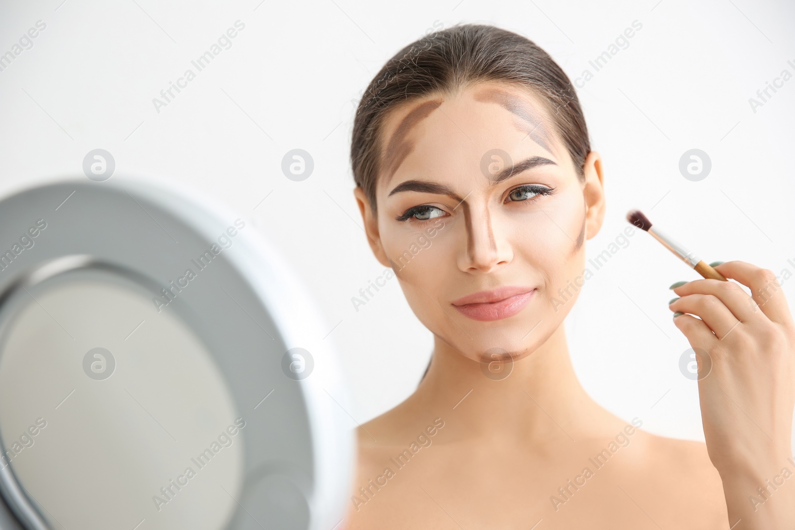 Photo of Young woman applying contouring lines on her face against light background. Professional makeup products