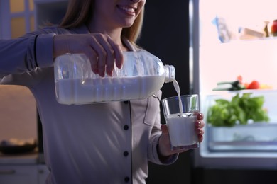 Young woman pouring milk from gallon bottle into glass near refrigerator in kitchen at night, closeup Photo of Young woman pouring milk from gallon bottle into glass near refrigerator in kitchen at night, closeup