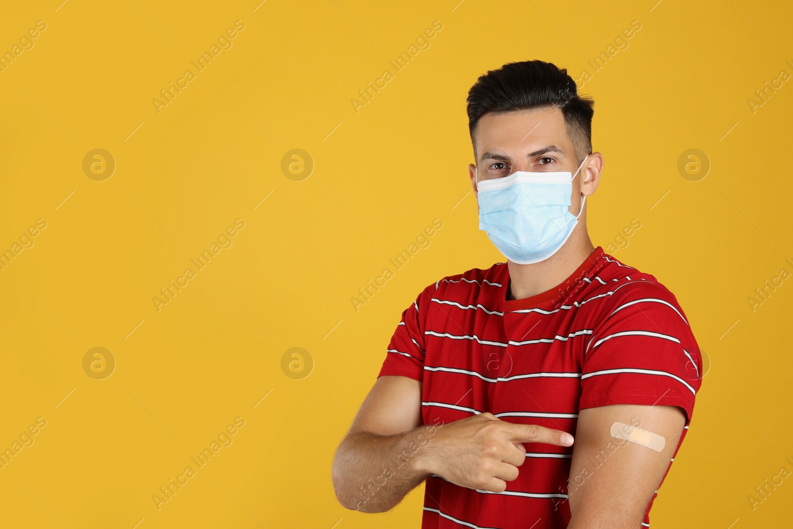 Vaccinated man with protective mask showing medical plaster on his arm against yellow background. Space for text Photo of Vaccinated man with protective mask showing medical plaster on his arm against yellow background. Space for text