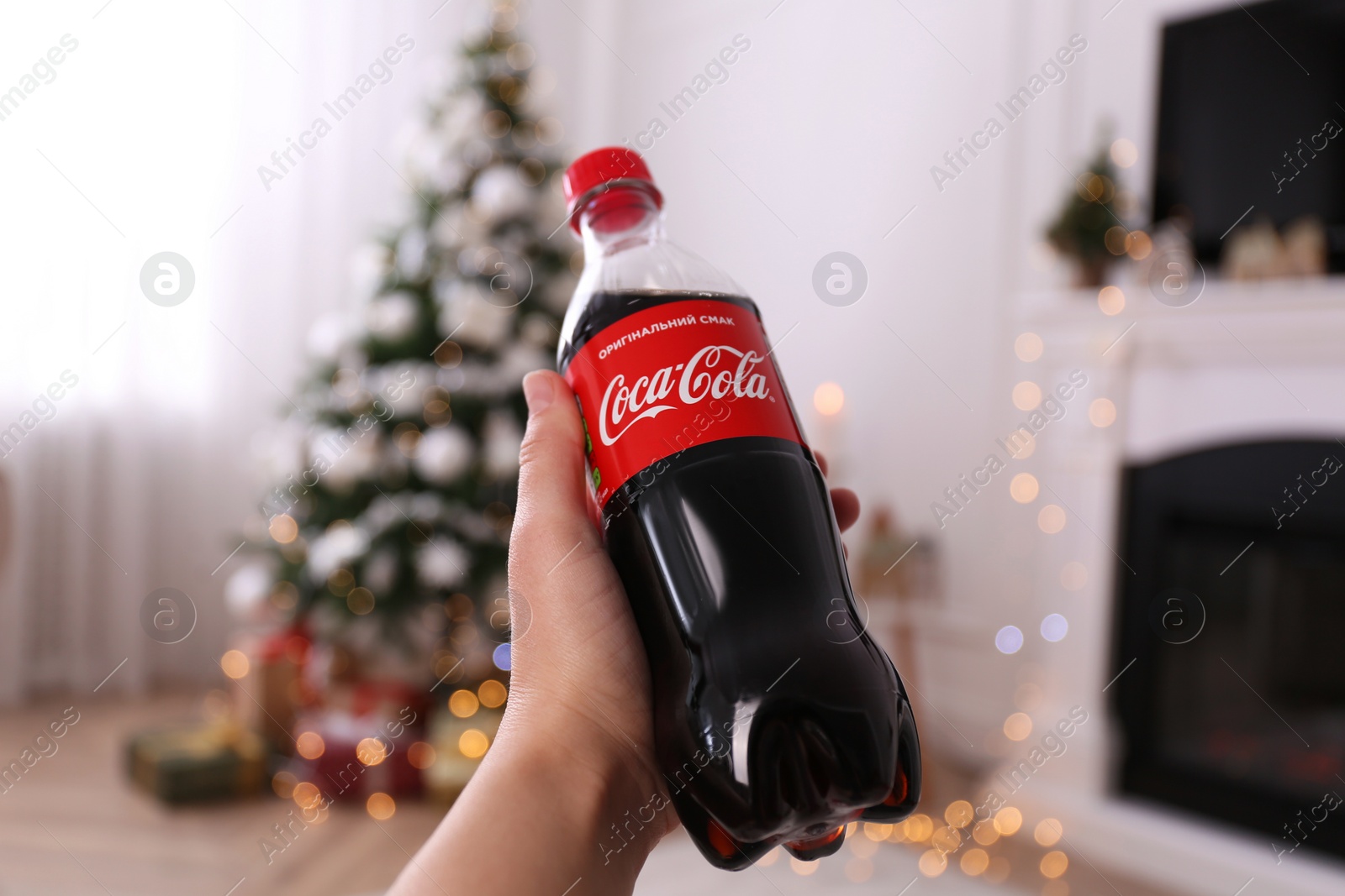 MYKOLAIV, UKRAINE - JANUARY 13, 2021: Woman holding bottle of Coca-Cola in room with Christmas tree, closeup Photo of MYKOLAIV, UKRAINE - JANUARY 13, 2021: Woman holding bottle of Coca-Cola in room with Christmas tree, closeup