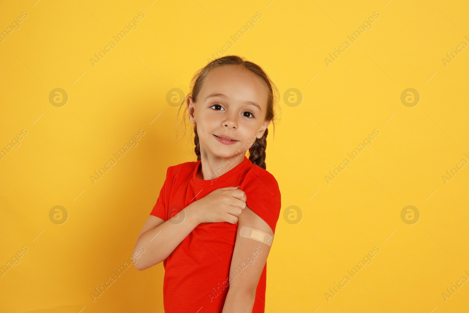 Vaccinated little girl showing medical plaster on her arm against yellow background Photo of Vaccinated little girl showing medical plaster on her arm against yellow background