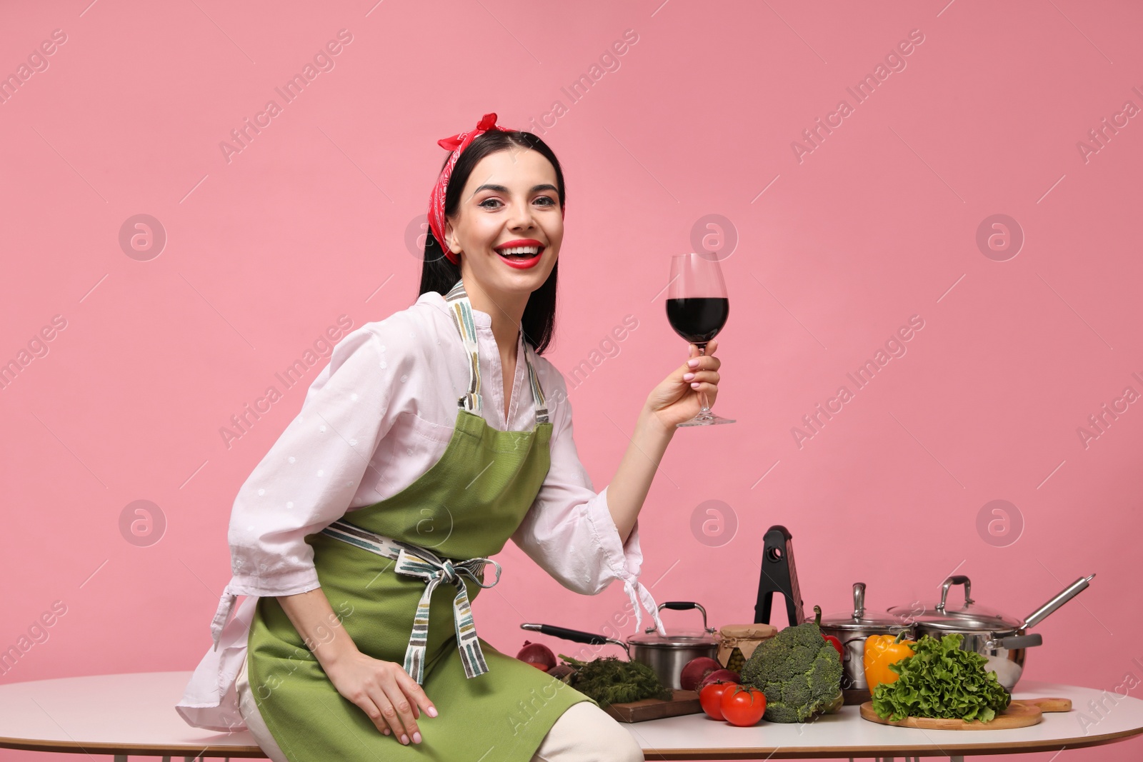 Young housewife with glass of wine, vegetables and different utensils on pink background Photo of Young housewife with glass of wine, vegetables and different utensils on pink background