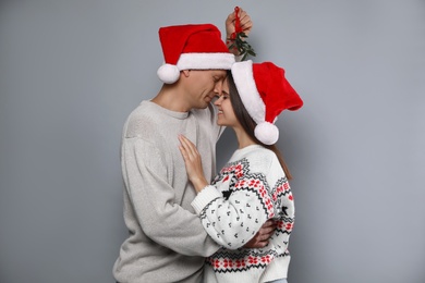 Happy couple in Santa hats standing under mistletoe bunch on grey background Photo of Happy couple in Santa hats standing under mistletoe bunch on grey background