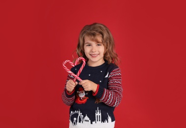 Cute little girl in Christmas sweater making heart shape with candy canes against red background Photo of Cute little girl in Christmas sweater making heart shape with candy canes against red background