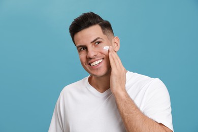 Happy handsome man applying face cream against turquoise background Photo of Happy handsome man applying face cream against turquoise background