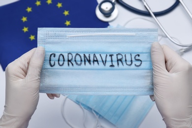 Doctor holding mask with word Coronavirus above medical items and European Union flag, closeup Photo of Doctor holding mask with word Coronavirus above medical items and European Union flag, closeup